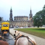 Im Vordergrund fahren zwei Pferdekutschen auf der Straße. Im Hintergrund mehrere Kirchen und die Semperoper in der Altstadt von Dresden