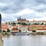 Eine Frau, links im Bild, schaut von der Karlsbrücke über die Moldau im Vordergrund auf das gegenüberliegende Ufer mit Teilen der Prager Altstadt, überragt durch die Anhöhe mit der Prager Burg