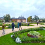 Einzelne Teilnehmer der Reisegruppe wandern über einen Weg zwischen Grünflächen mit Blumenbeeten vor einem barocken Gebäudeteil der Schlosses Pillnitz