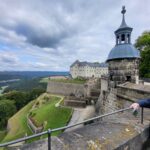 Ein Mann, recht im Vordergrund schaut von der Festungsbrüstung auf die Elbe am Linken Bildrand hinab. Im Hintergrund des Mannes ein Wachtturm und Festungsgebäude hinter der Festungsmauer am steilen Abhang der Festung.