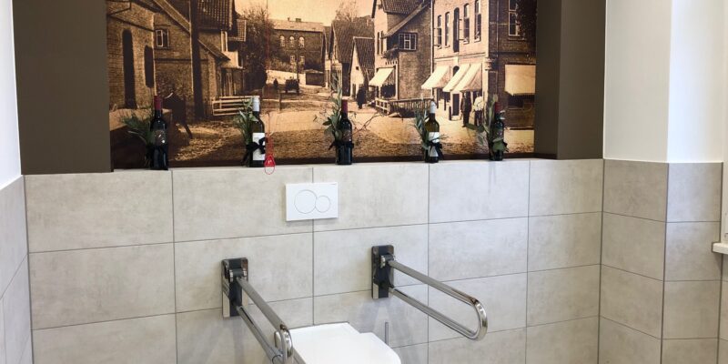 Sanitärraum mit barrierefreier Toilette inklusive Haltegriffen und Notfall-Zugschnur. Im Hintergrund an der Wand eine historische Ansicht der Marktstraße in Harsefeld.